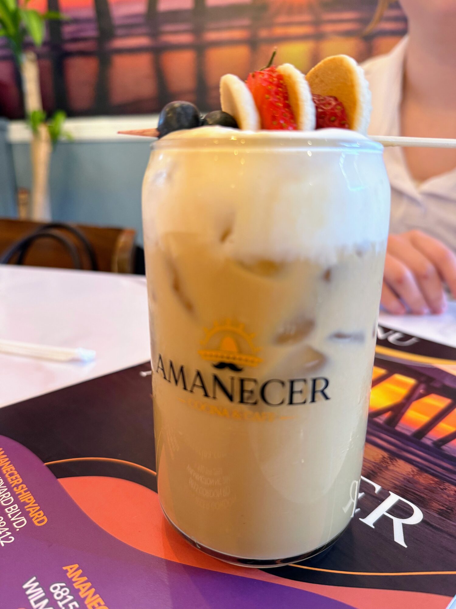 A tall iced horchata-style latte in an Amanecer-branded glass, garnished with strawberry, banana and blueberries.