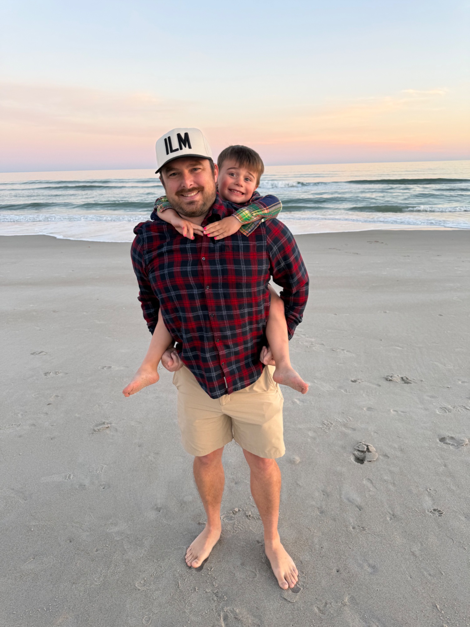 An adult in an ILM cap giving a child a piggyback ride on the beach at sunset near Wilmington, NC.