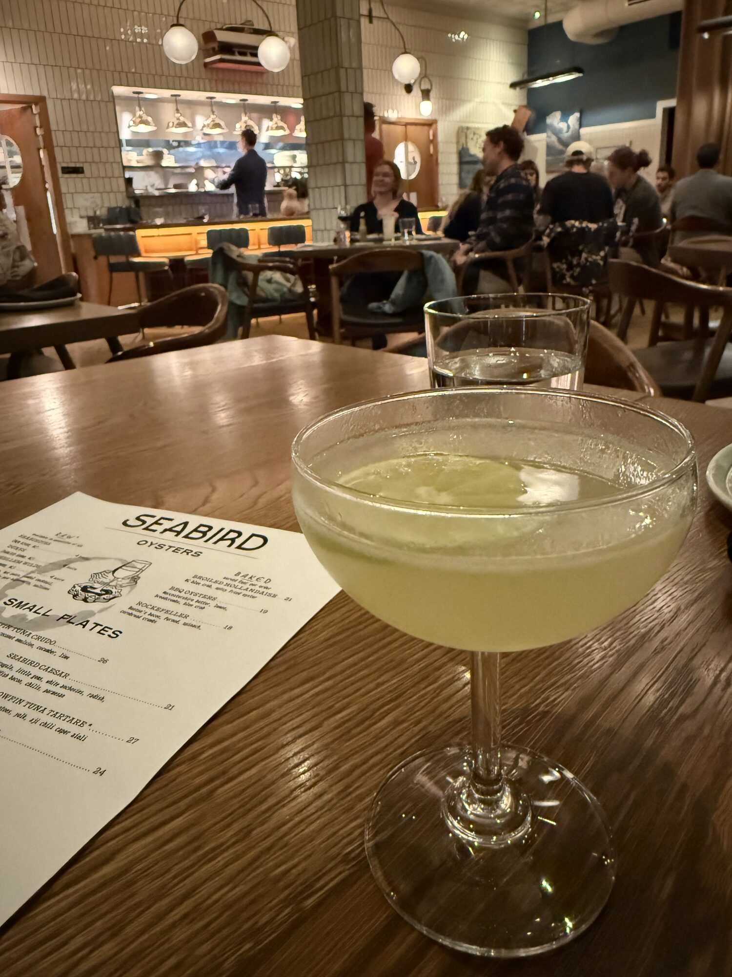 A pale green cocktail in a coupe glass beside a Seabird oysters and small plates menu, with the open kitchen behind.